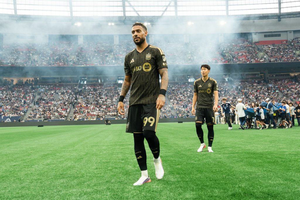 Two soccer players in black-gold jerseys walk onto a green pitch as smoky stadium lights rise and a packed arena watches.